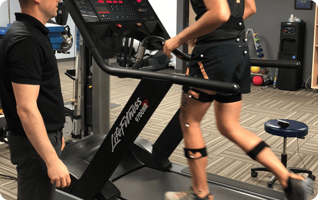 A man observing a gait analysis session, focused on a person walking on a treadmill with motion-capture markers attached. The man is using a monitor to assess walking patterns, stride, and posture for evaluation and potential improvement.